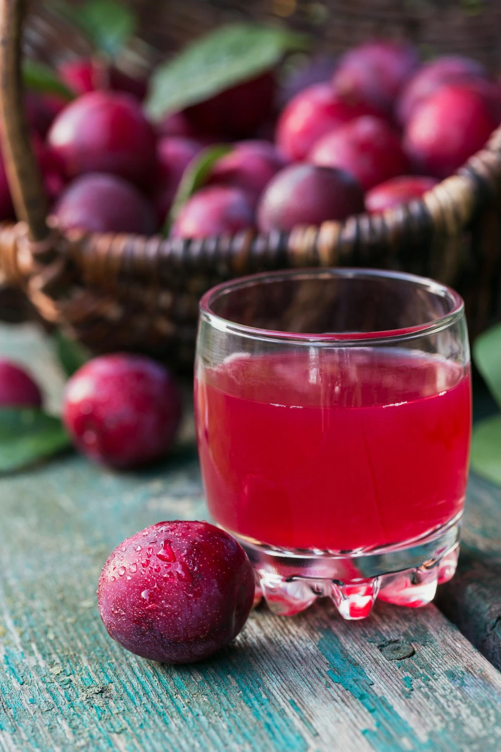 A glass of fresh plum juice with ripe plums and mint leaves on a wooden table.