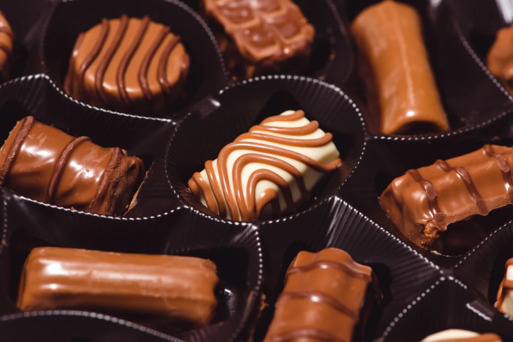 Pouring melted chocolate into silicone molds with bowls of ingredients on a marble counter.